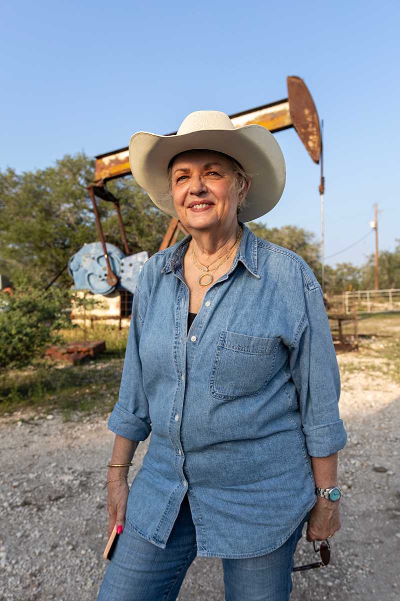 Marsha in denim shirt, jeans, and cowboy hat standing in front of an oil pumper