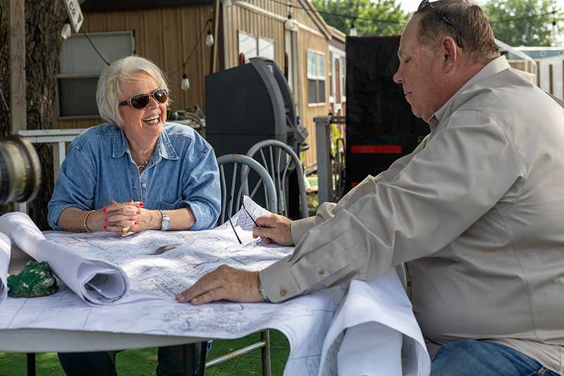 Marsha in denim shirt and black sunglasses talking with a man in a tan shirt as they look at drilling plans spread out on a table